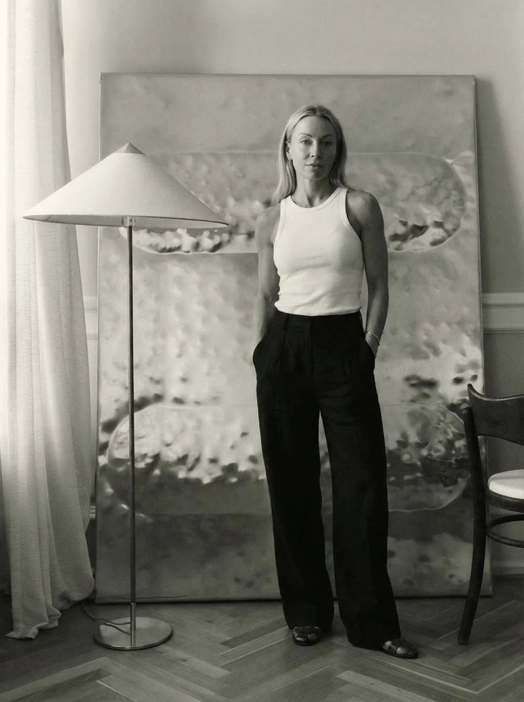 Black and white photo of a woman with long hair wearing a sleeveless top and black pants, standing next to a floor lamp, in front of a large abstract painting, with a chair to the right.