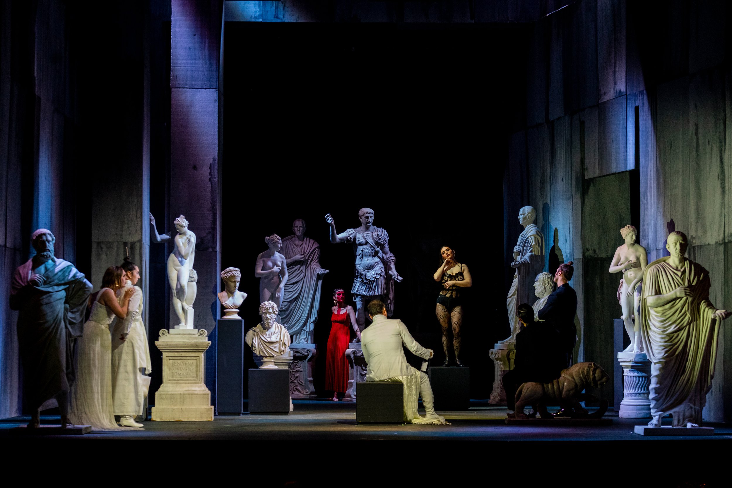 A theatrical stage set resembling ancient Greek or Roman statues, with actors and actresses performing amid classical sculptures and busts, some seated and others standing, under dramatic lighting.