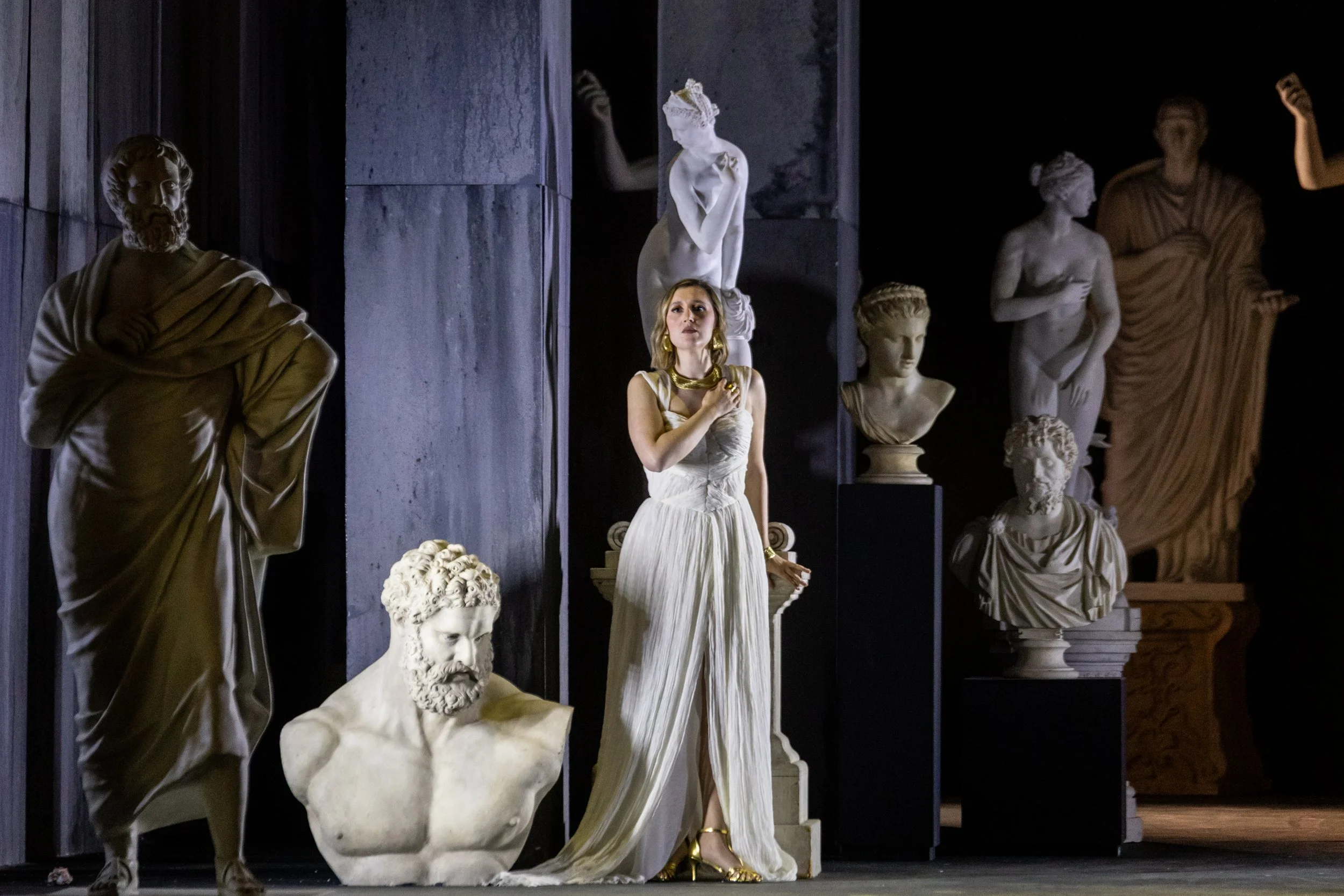 Female opera performer in white stands among classical statues in a sculptural stage setting in Abos’ Pelopida