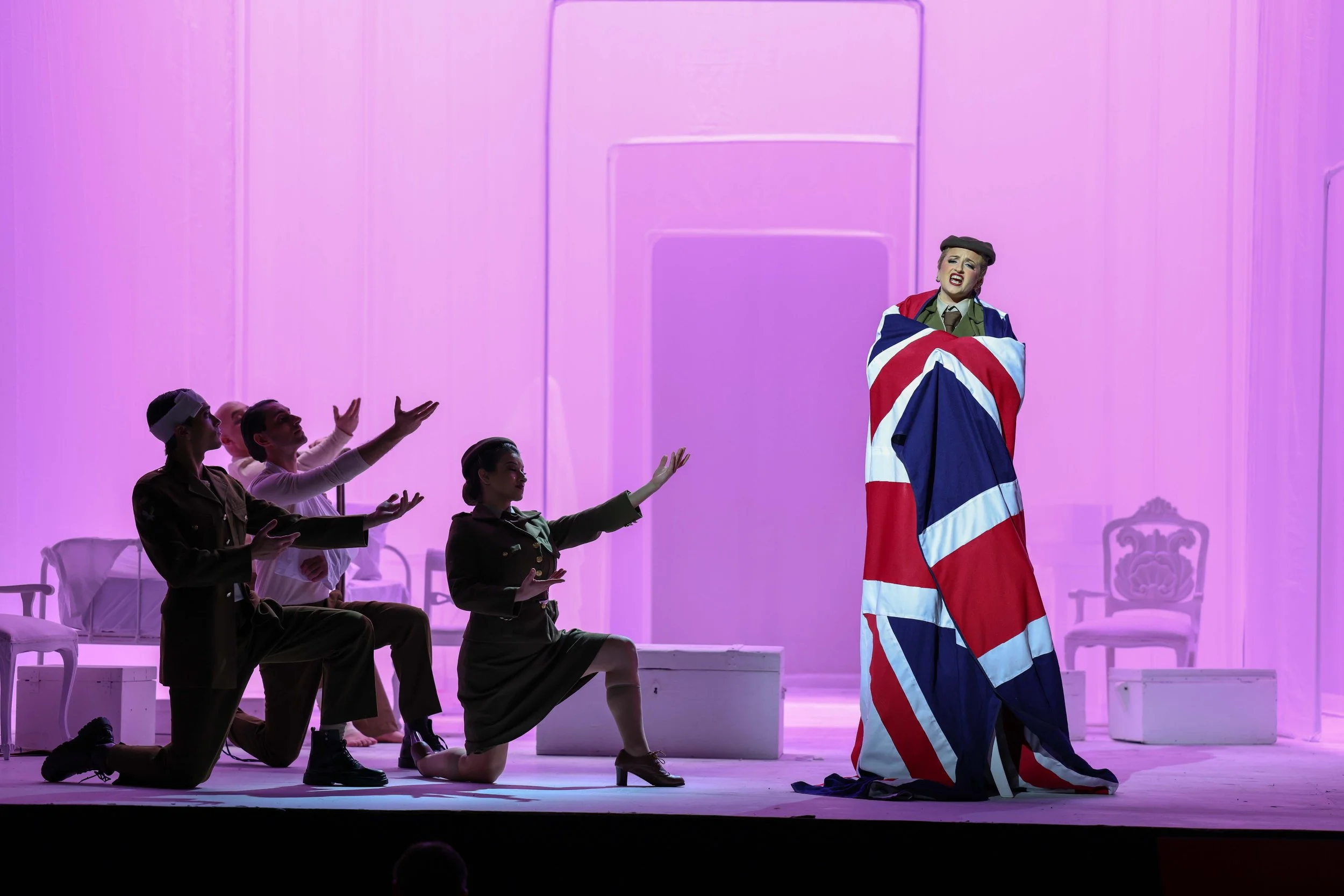 Scene from Gluck’s La corona at the Valletta Baroque Festival, directed by Brett Nicholas Brown, featuring a central performer draped in a Union Jack flag with surrounding figures in a 1940s wartime setting