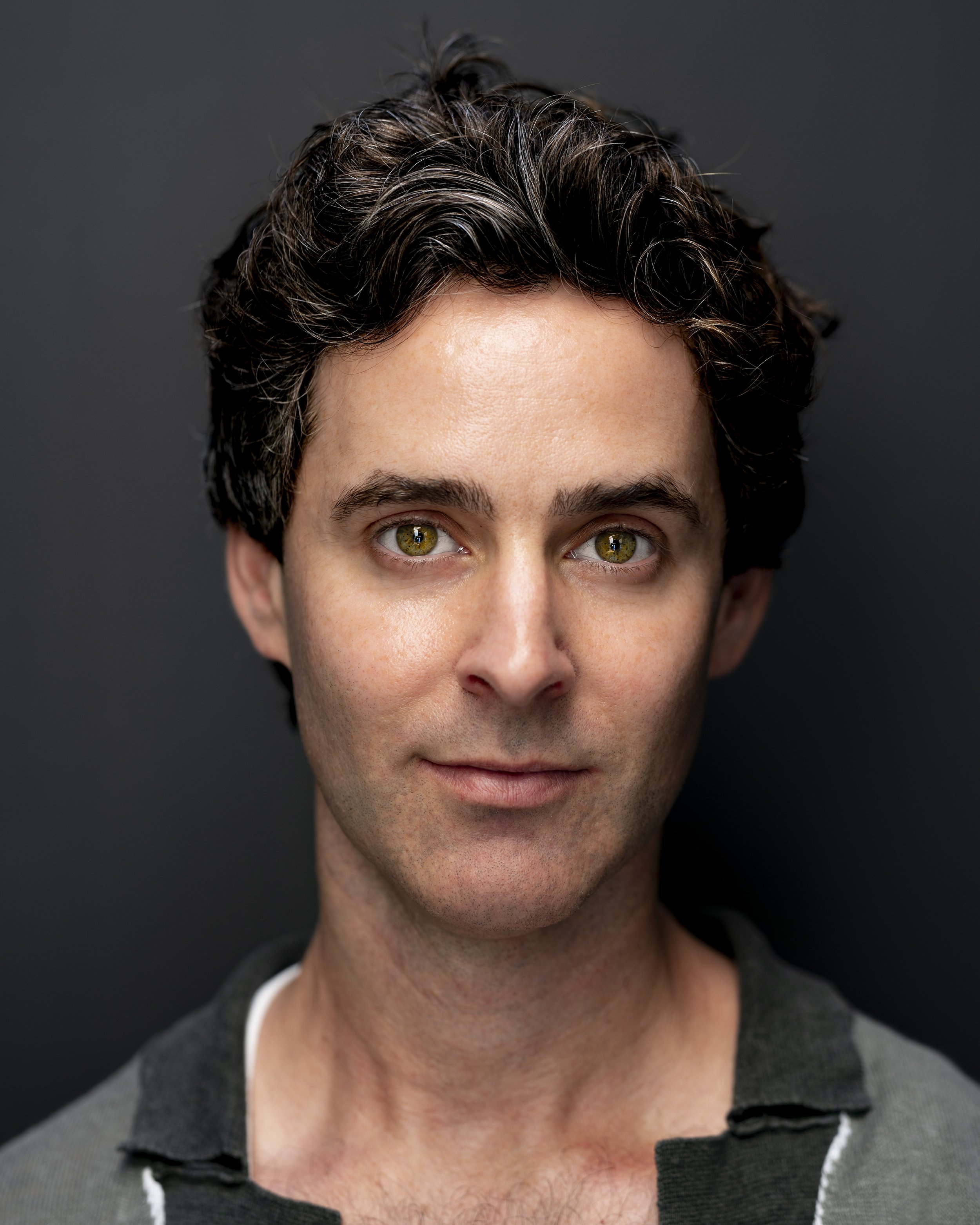 Headshot of Brett Nicholas Brown. He has dark curly hair and green eyes, and wears a green shirt with a dark collar, set against a dark background.