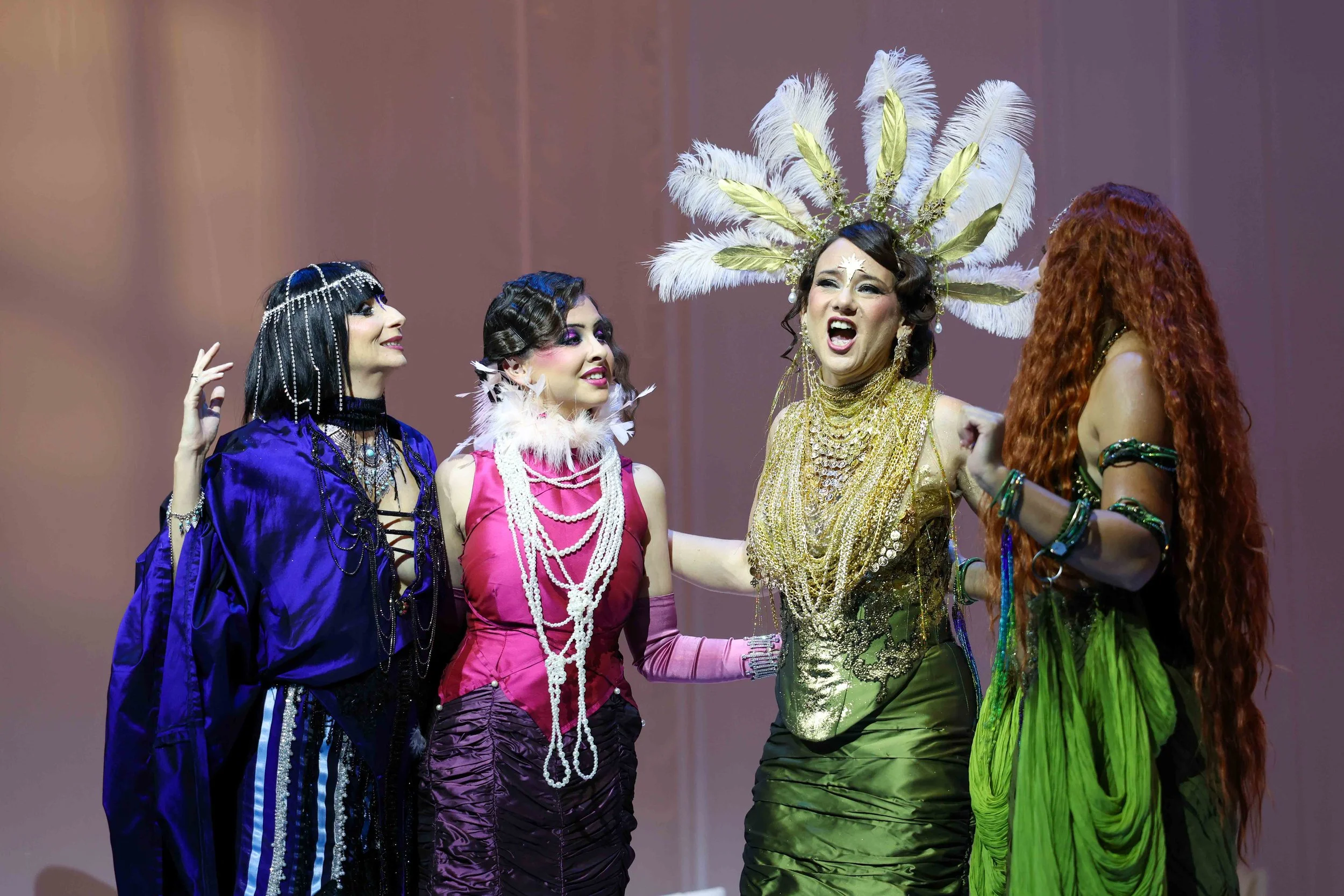 Four women dressed in colorful, elaborate costumes engaged in a theatrical performance. One woman wears a gold dress with an ornate feathered headdress, another in a pink and purple outfit with feathers and pearl necklaces, a third in a shiny purplish dress with layered jewelry, and the last in a green skirt with long, curly red hair.