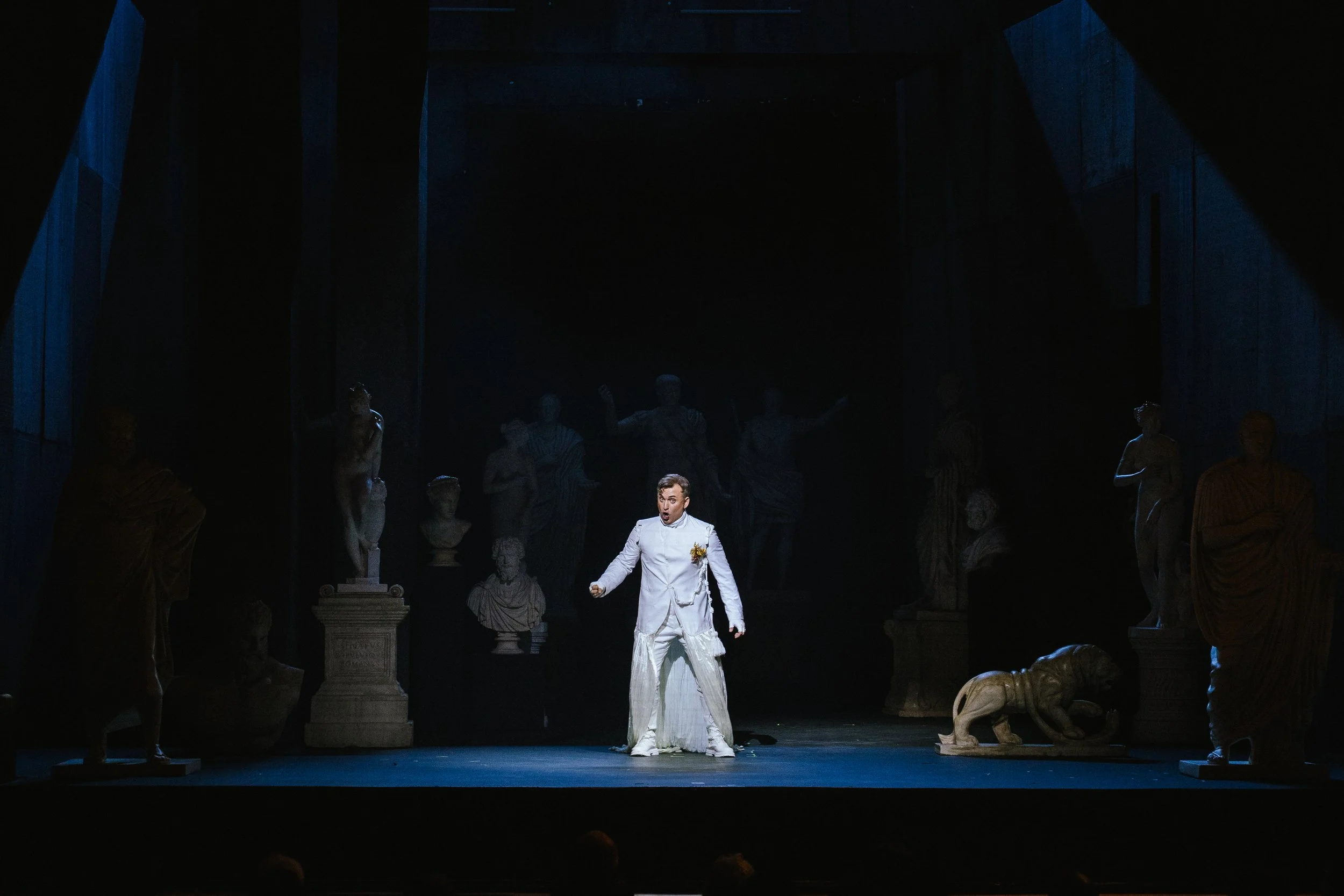 Male opera performer in white costume stands alone among classical statues on a dark stage in Abos’ Pelopida, directed by Brett Nicholas Brown