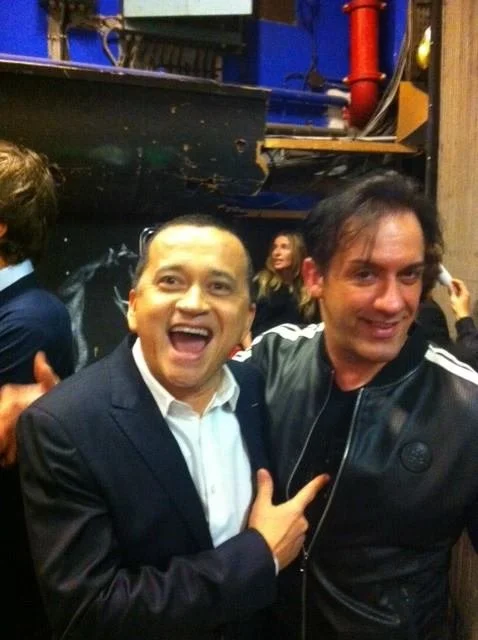 Deux hommes souriants posant pour une photo dans un environnement intérieur, avec d'autres personnes en arrière-plan.