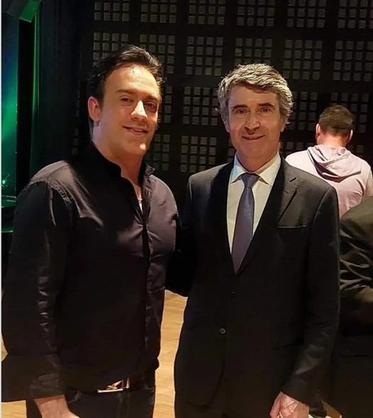 Deux hommes posent pour une photo, l'un en tenue décontractée avec une chemise noire, l'autre en costume avec cravate, dans un environnement intérieur