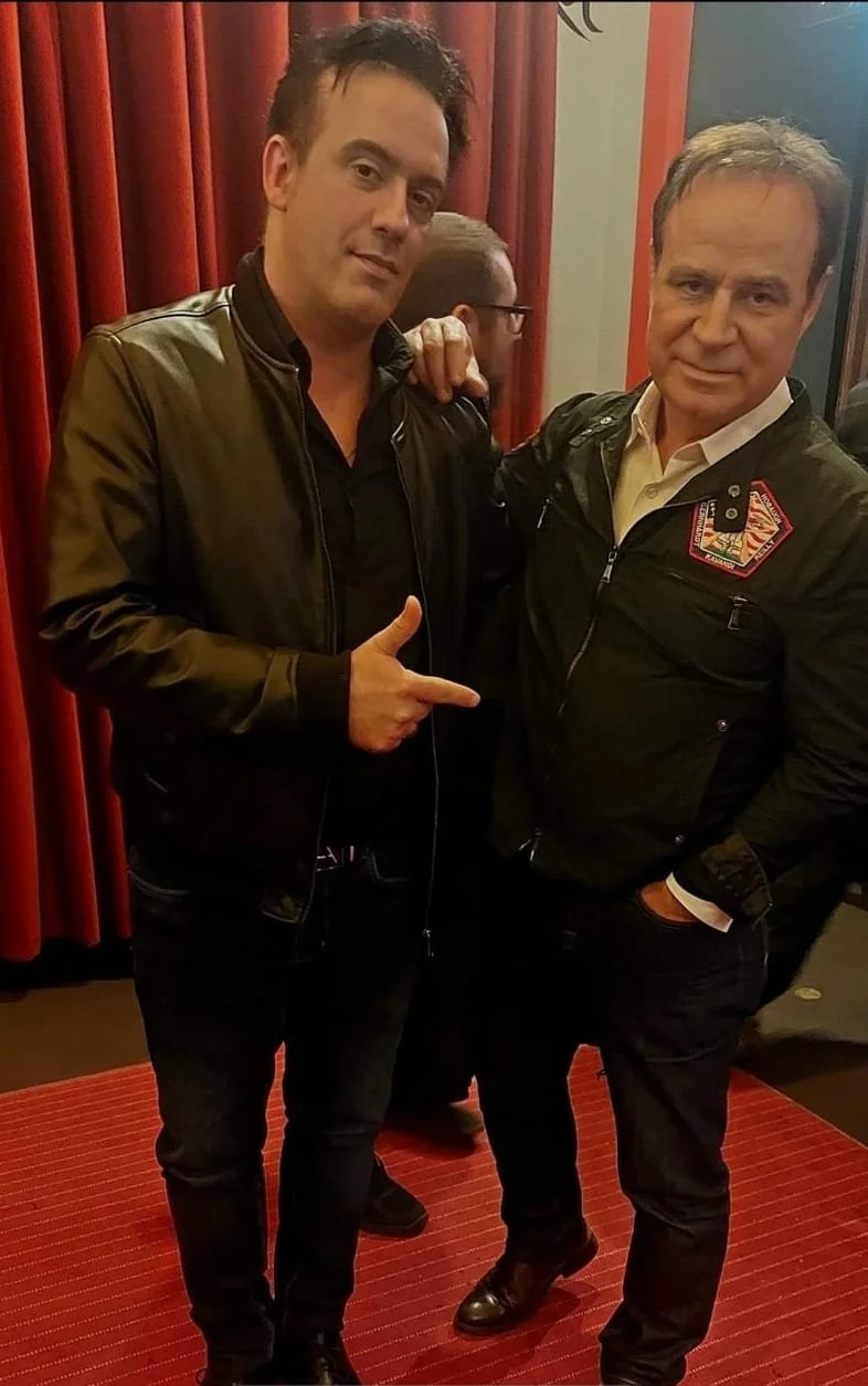 Deux hommes posent pour une photo en intérieur, l'un portant une veste en cuir noire et pointant du doigt vers l'autre, qui porte une veste avec un patch sur la poitrine. En arrière-plan, il y a une personne portant des lunettes et des rideaux rouges