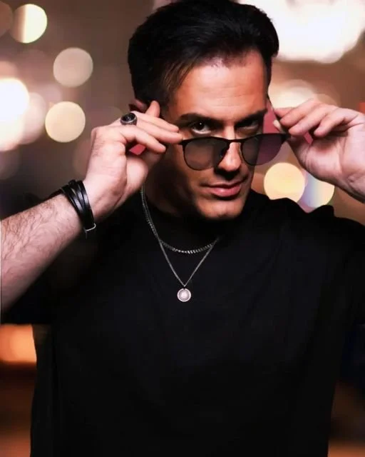 Un homme portant des lunettes de soleil, un t-shirt noir et un collier, ajustant ses lunettes dans un décor avec des lumières floues en arrière-plan.