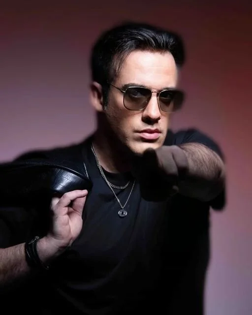 Un homme portant des lunettes de soleil noires et un t-shirt noir, avec une veste à l'épaule et un collier, pose avec un poing levé devant un fond flou.