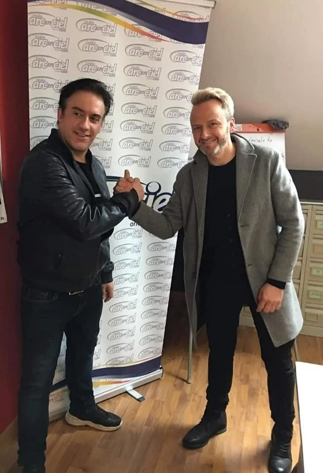 Deux hommes se serrent la main et sourient, devant un fond avec le logo 'arc-en-ciel'.