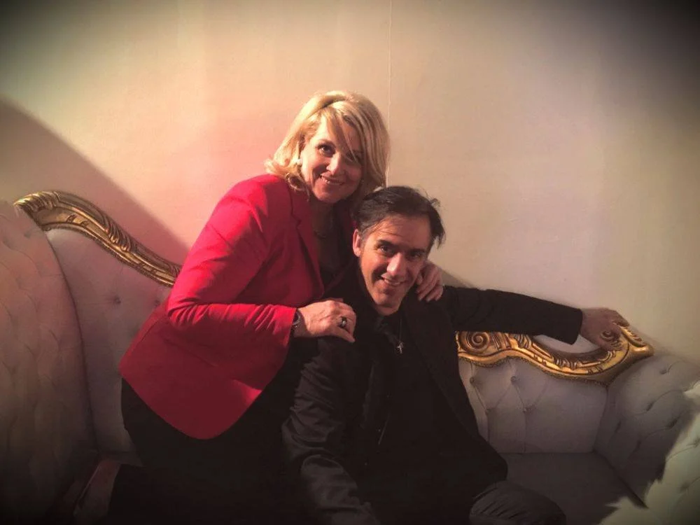 Une femme blonde en veste rouge se tient près d’un homme assis sur un canapé, tous deux souriants.