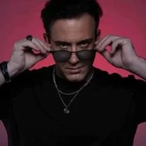 Un homme portant des lunettes noires, tenant ses lunettes avec les mains, avec un fond rose.