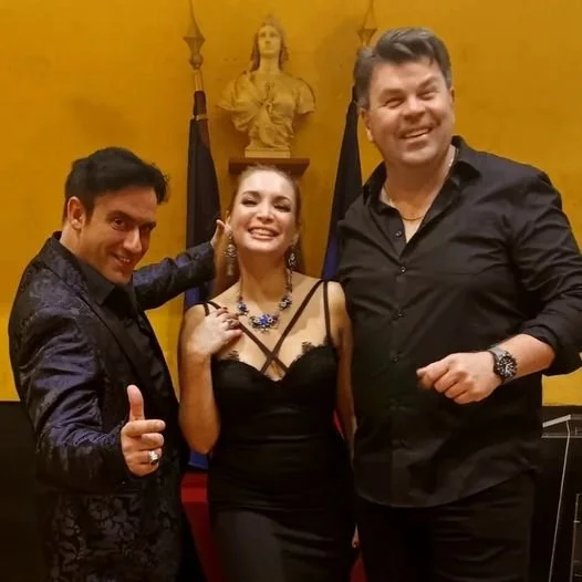 Trois personnes souriantes posent devant un décor jaune avec une statue en arrière-plan lors d'une célébration ou d'un événement formel.