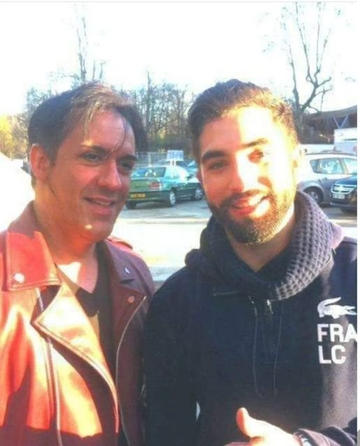 Deux hommes posant pour une photo à l'extérieur, avec des voitures en arrière-plan.