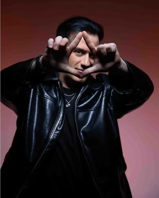 Jeune homme regardant l'objectif, portant une veste en cuir noire, faisant un symbole avec ses mains en formant un triangle, fond neutre