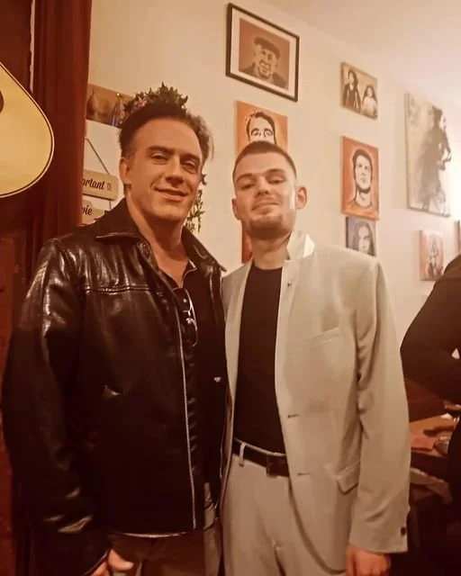 Deux hommes posent pour une photo dans une pièce décorée de portraits encadrés au mur.