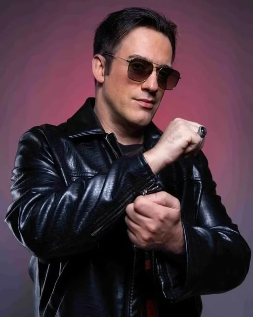 Un homme portant des lunettes de soleil et un blouson en cuir noir pose avec un attitude confiante, main levée en poing.