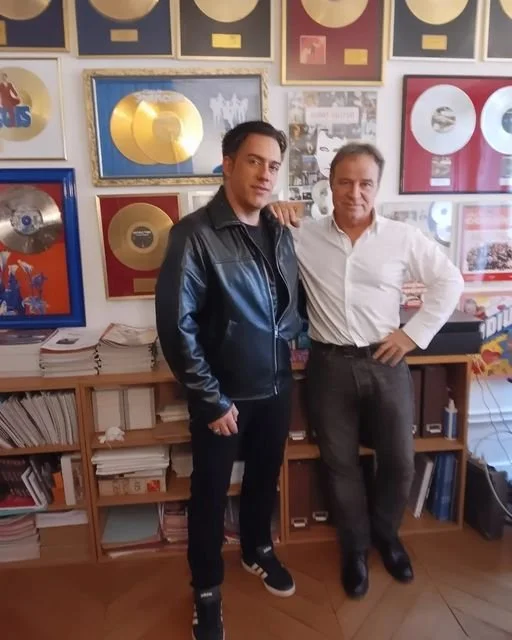 Deux hommes posent ensemble dans une pièce décorée avec de nombreux disques d'or et d'argent encadrés aux murs, à côté de meubles remplis de piles de livres et papiers.