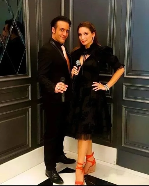 Un homme et une femme élégamment habillés posent ensemble dans un mur en bois foncé, tenant des microphones, la femme porte des chaussures à talons rouges.