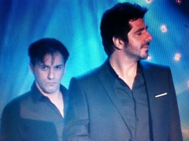 Deux hommes en costume noir sur un fond avec des lumières bleues et rouges.