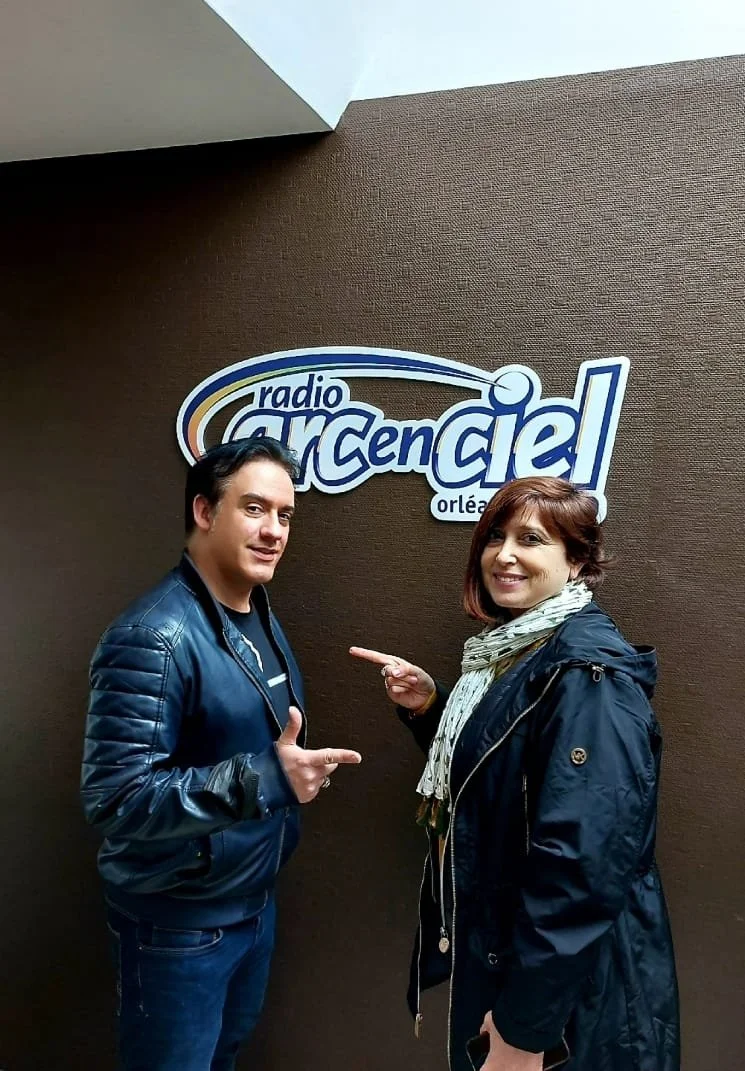 Deux personnes pointant vers un logo de Radio Arcenciel à Orléans, devant un mur brun. Personne à gauche portant une veste en cuir bleue, personne à droite avec une veste noire et une écharpe, souriantes.