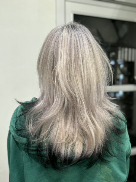 Back of a person with shoulder-length blonde hair with darker tips, wearing a green hoodie, standing indoors near a window or door.