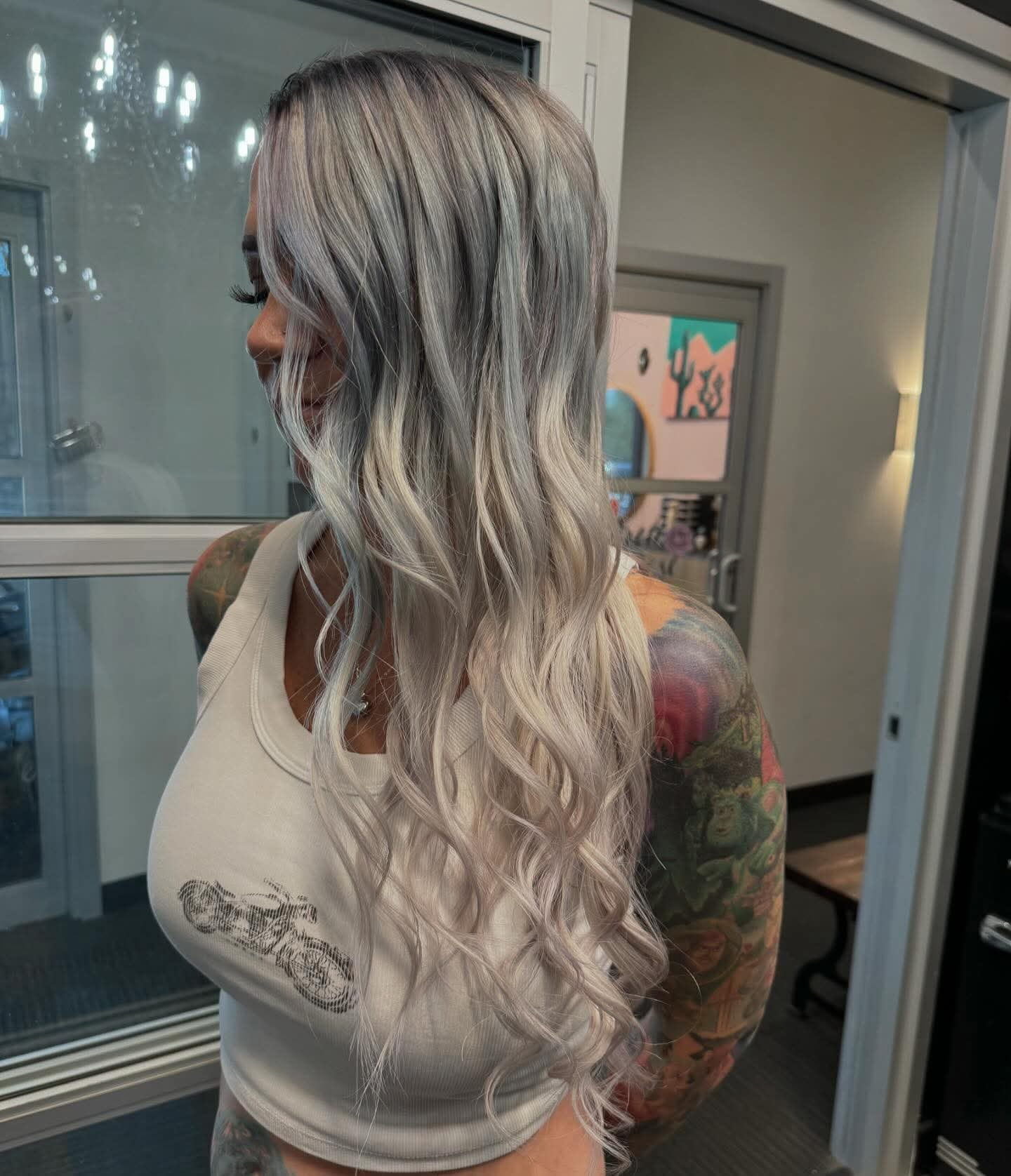 A woman with long, wavy, bleach-blonde hair and tattoos on her arms, wearing a white tank top, standing indoors near a glass door.