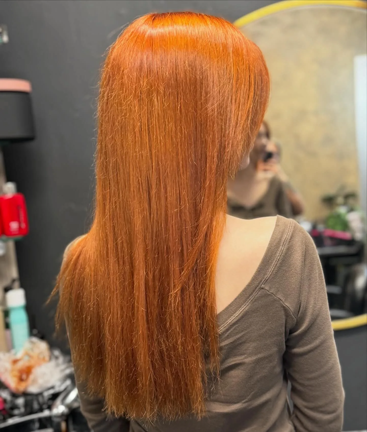 Person with long, straight, vibrant orange hair, wearing a brown top, in a salon setting.