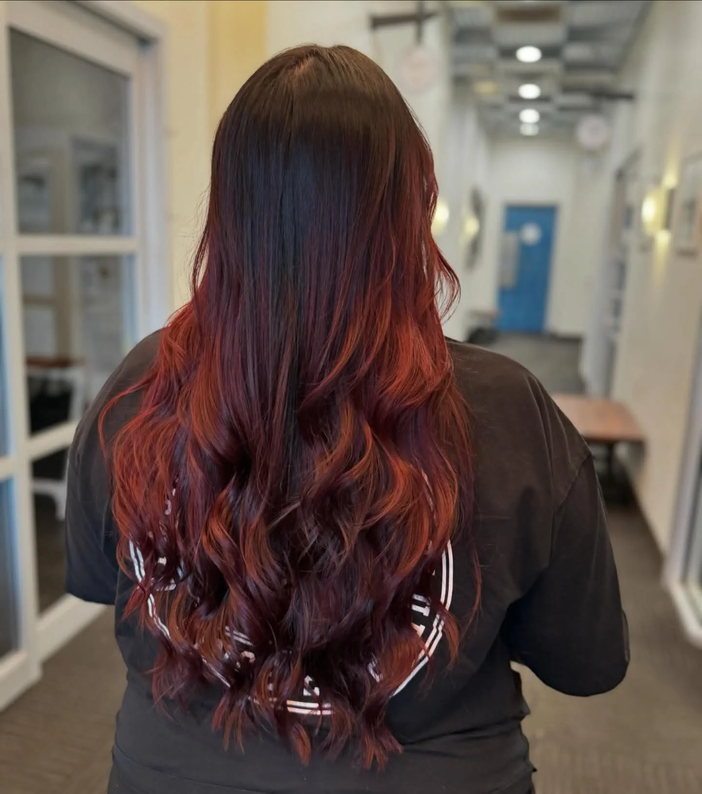 Back view of a woman with long, wavy hair dyed in shades of red and black, standing in a hallway.