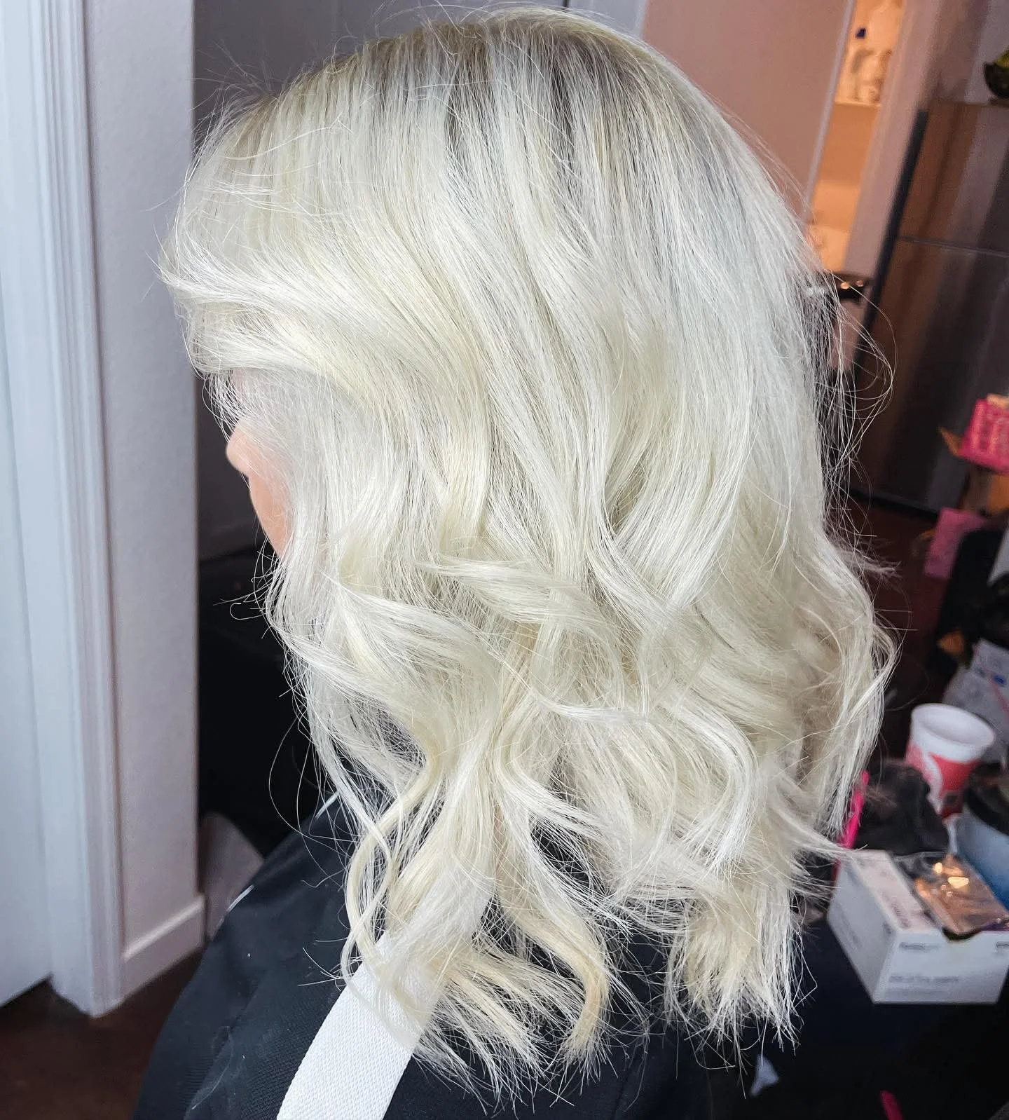 Blonde, curly hair styled in loose waves. The background shows a room with various items on surfaces and a doorway.