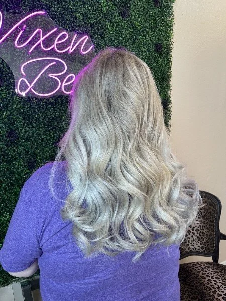 Back view of a woman with long, wavy blonde hair, wearing a purple shirt, standing in front of a green leafy wall with a pink neon sign that says 'Luxen B' and a leopard print chair nearby.