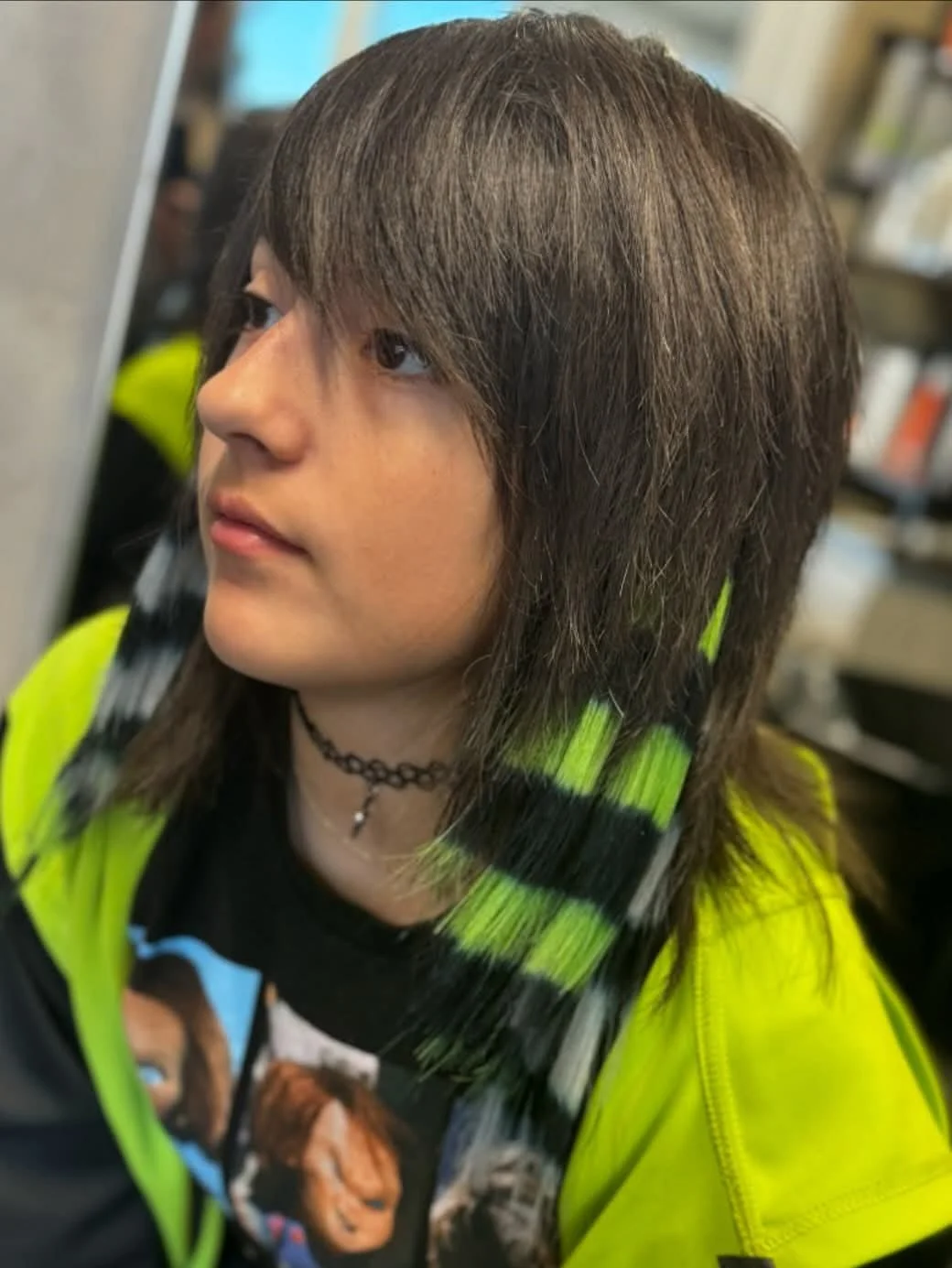 A young person with shoulder-length brown hair getting a haircut in a salon, wearing a bright yellow stylist coat and a black graphic t-shirt.