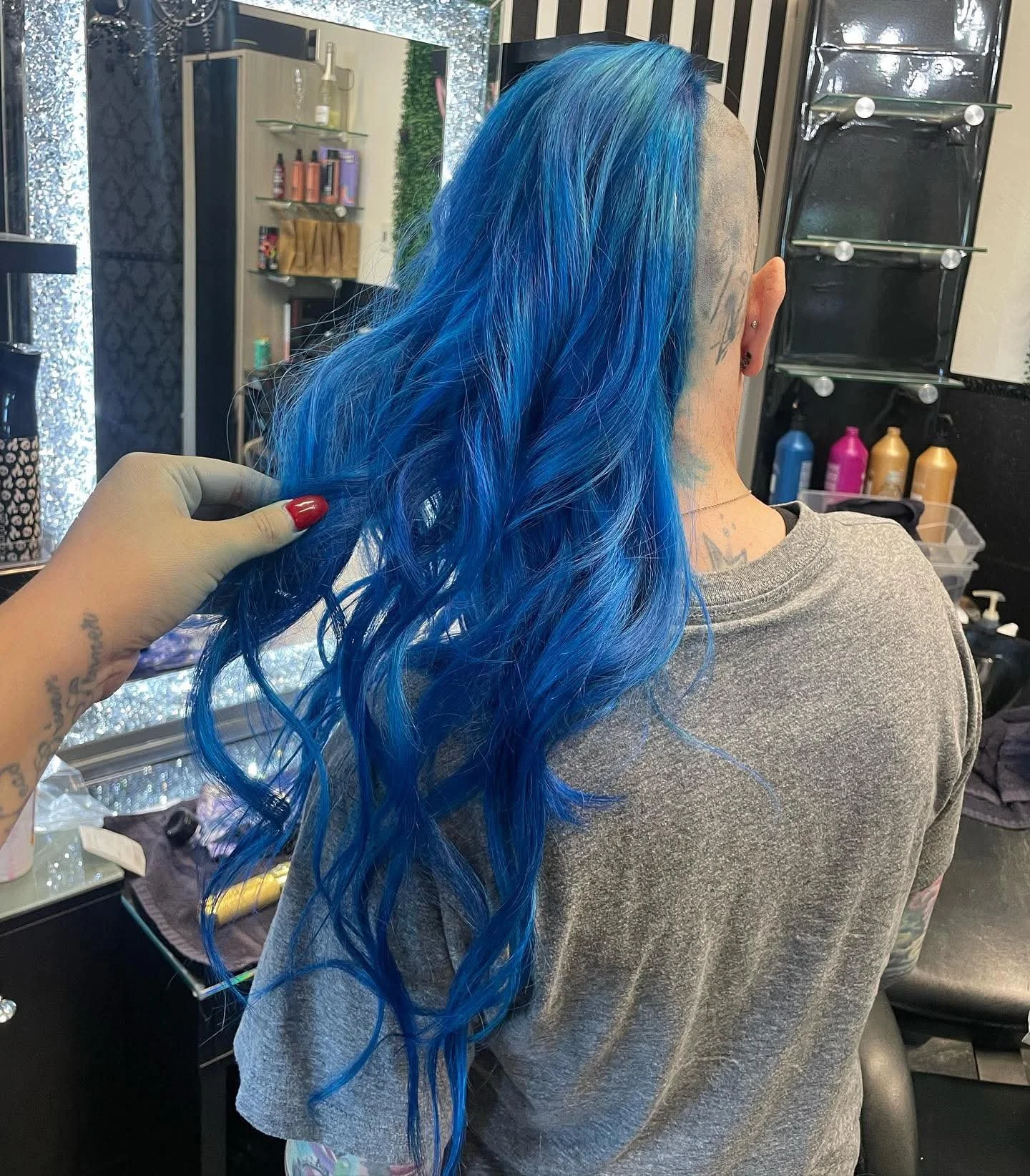 A person with long, bright blue, wavy hair sitting in a hair salon. Another person, with a tattoo on their hand and red nail polish, is holding and touching the hair. The salon has various hair products on shelves and bottles on a counter.