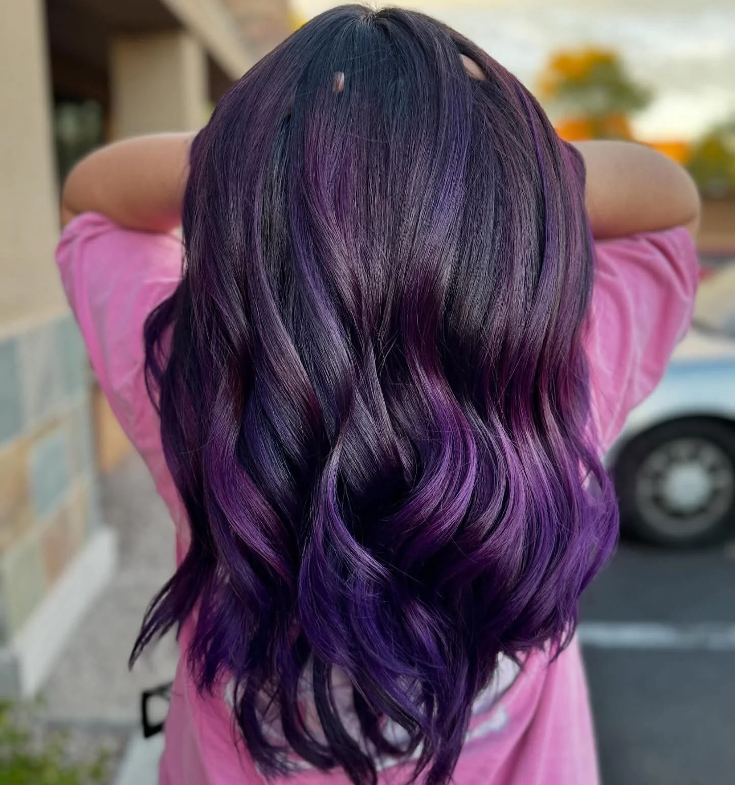 Person with wavy black hair dyed purple at the ends, wearing a pink shirt, standing outdoors with blurred background