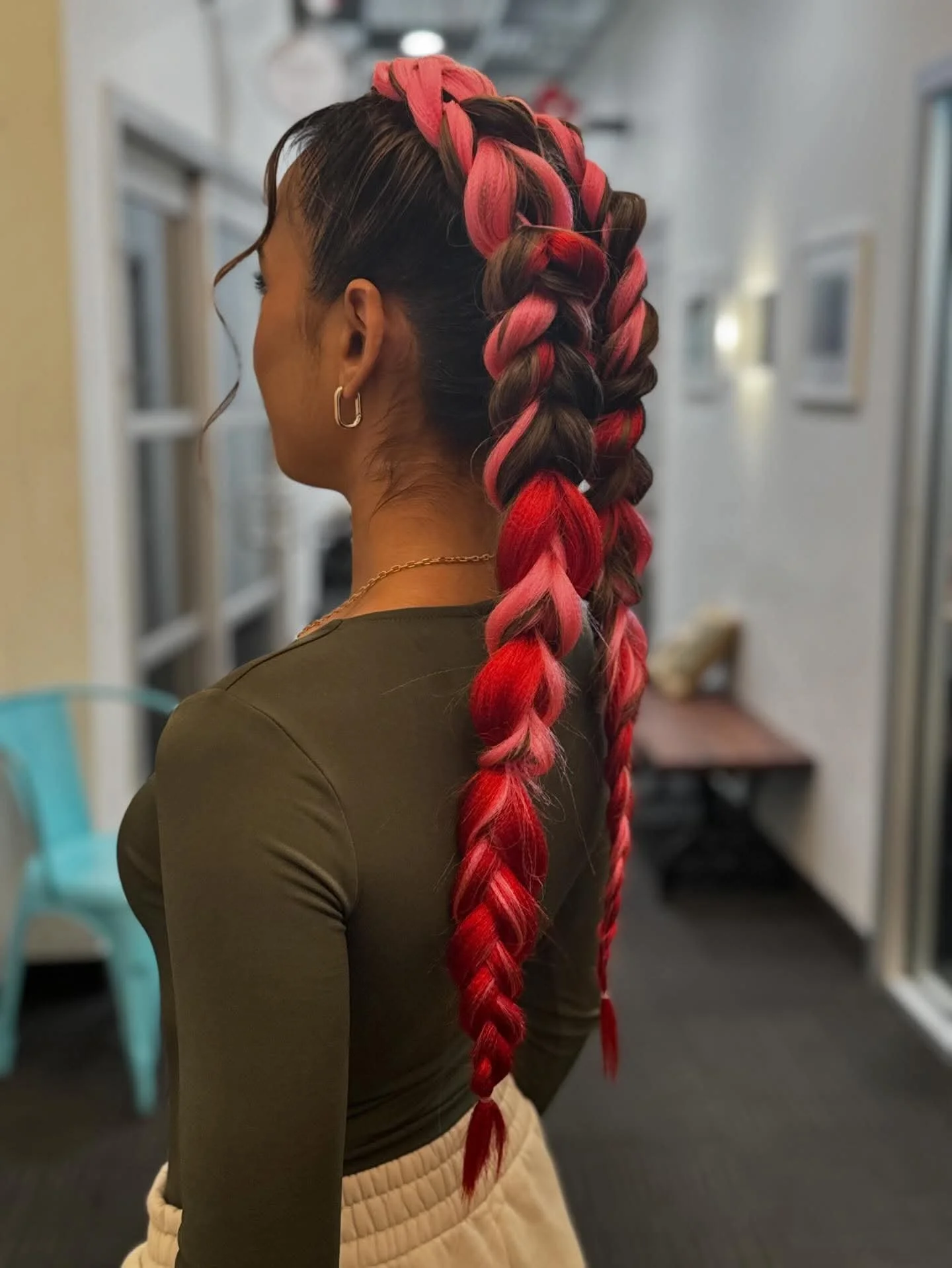 A woman with long, thick hair styled in a high ponytail with large, colorful braids in shades of red and pink, standing indoors with blurred background.