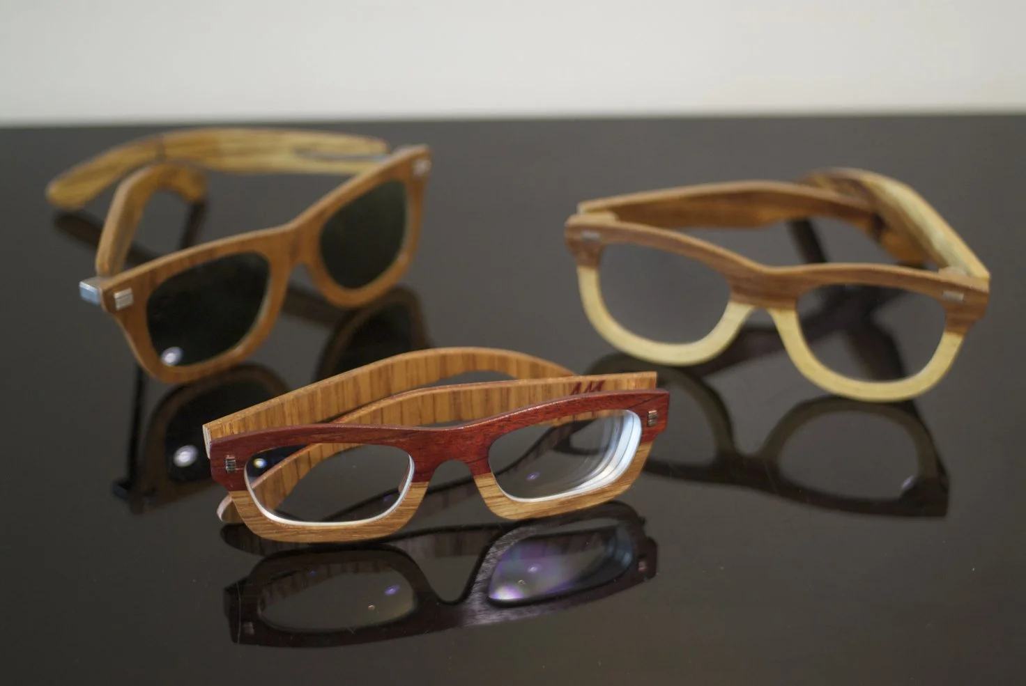 Three pairs of wooden sunglasses on a reflective black surface, with different wood finishes and dark lenses.