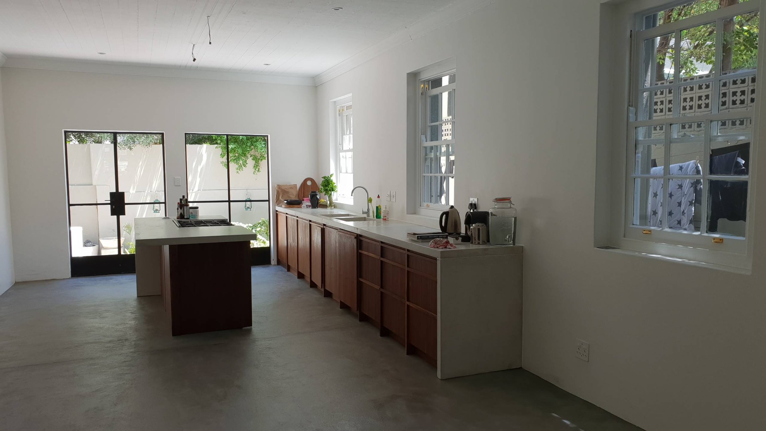 Empty kitchen with white walls, wooden cabinets, a white countertop, and windows letting in natural light. Items like a kettle, water filter, and some bottles are on the counter. Outside the windows, greenery and laundry hanging are visible.