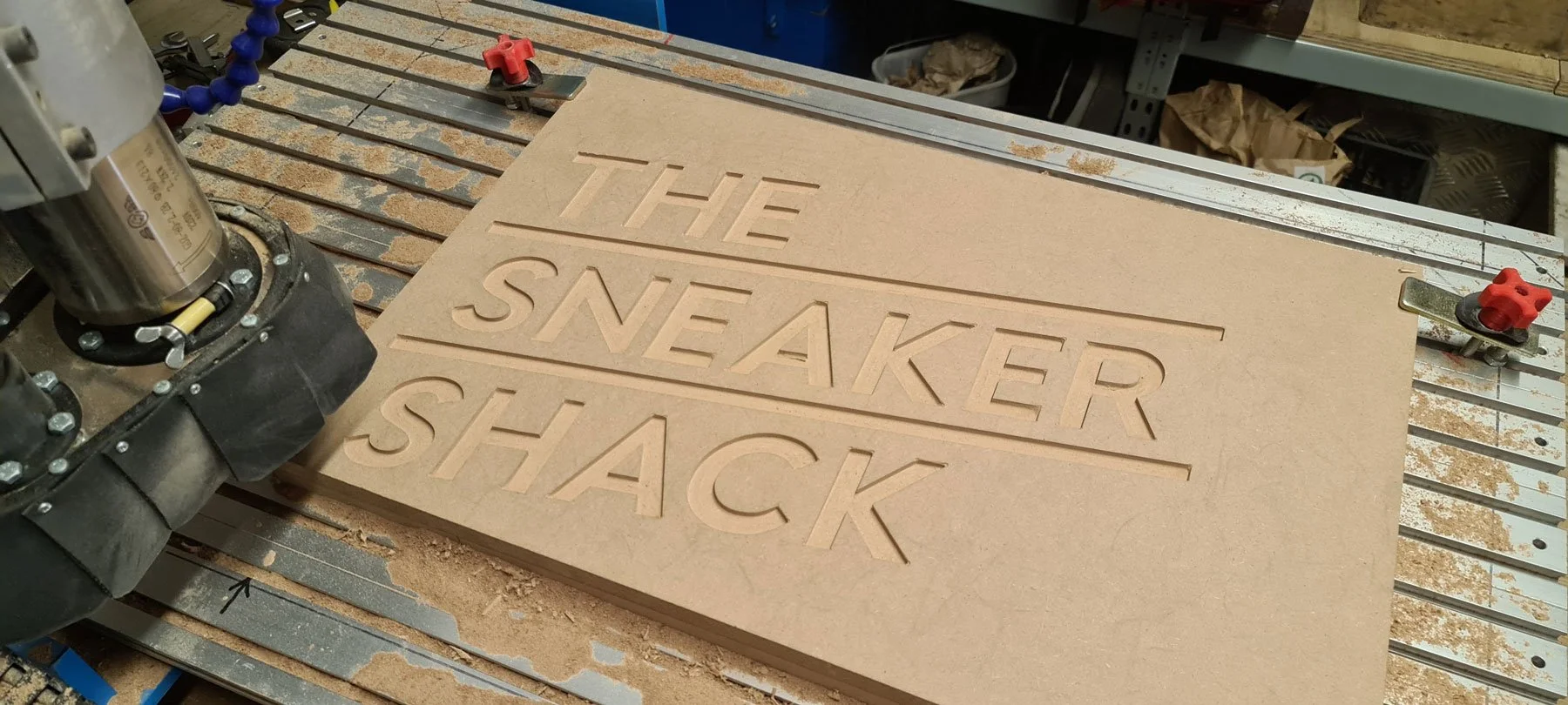 Wooden sign with the text 'THE SNEAKER SHACK' cut out, being shaped on a CNC machine in a woodworking shop.