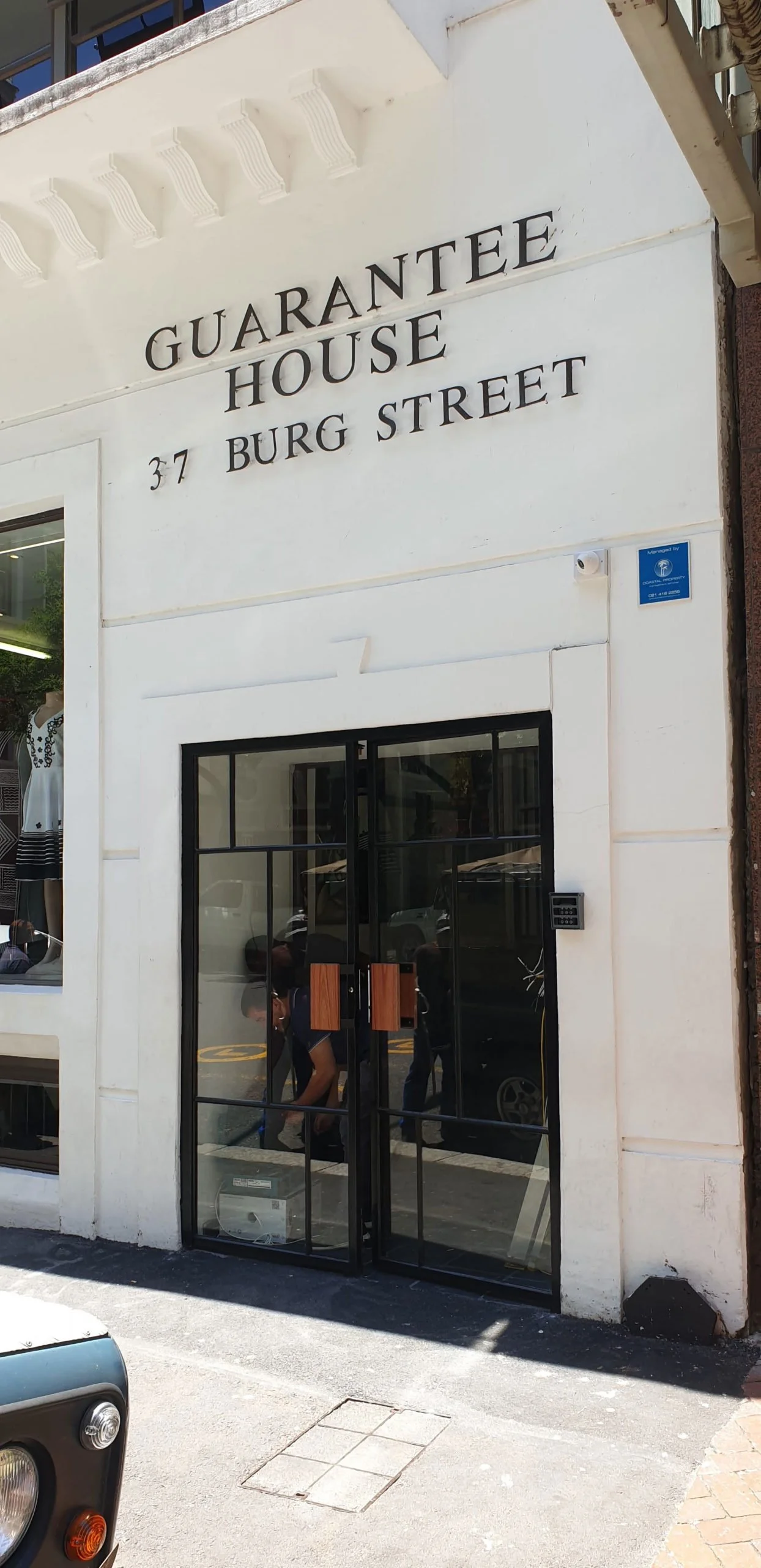 White building with black sign reading "Guarantee House 37 Burg Street" on the facade, and glass door with black metal frame.