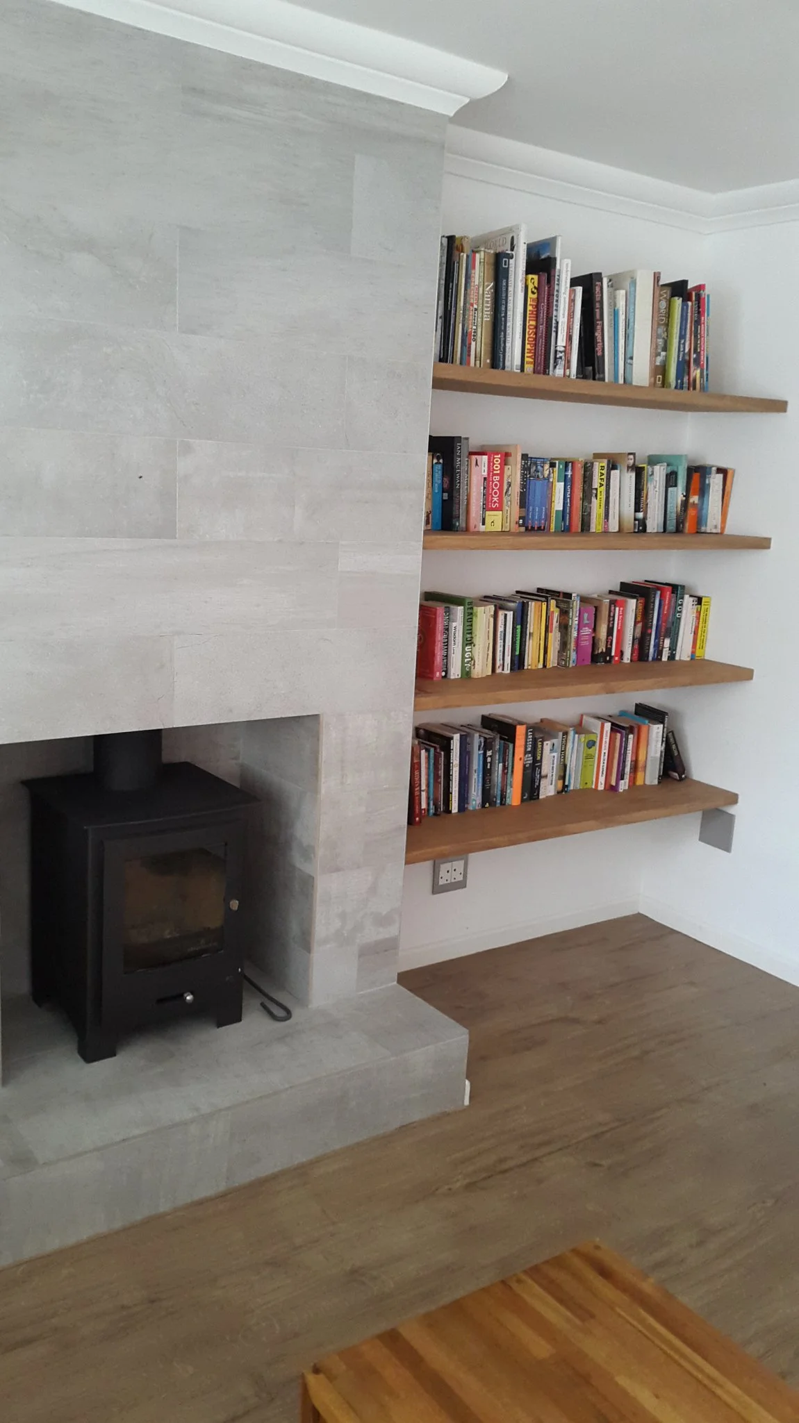 Living room corner with a wood-burning stove set into a grey stone surround, and three wooden floating bookshelves filled with books on a white wall.