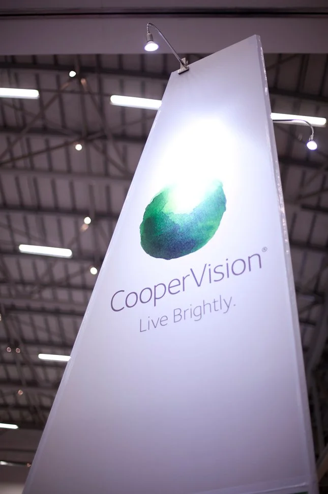 An illuminated vertical banner displaying the CooperVision logo with a green and blue stylized eye and the tagline 'Live Brightly,' set inside a large indoor space with a high ceiling and industrial lighting.