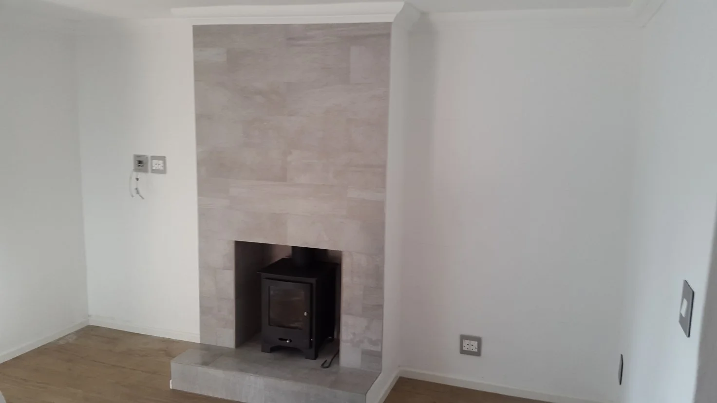 Empty living room corner with fireplace, beige tile surrounding the fireplace, wood flooring, electrical outlets, and white walls.