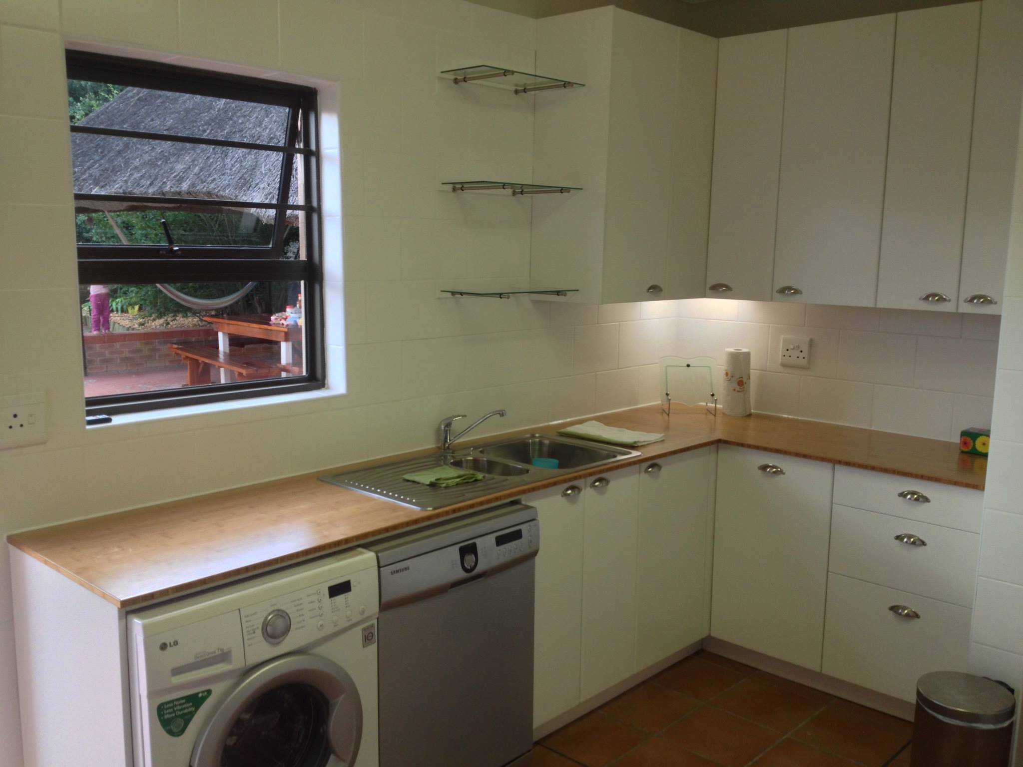 A kitchen with white cabinets, a wooden countertop, a stainless steel sink, a washing machine, a dishwasher, and a window showing an outdoor picnic table with a thatched roof and trees.
