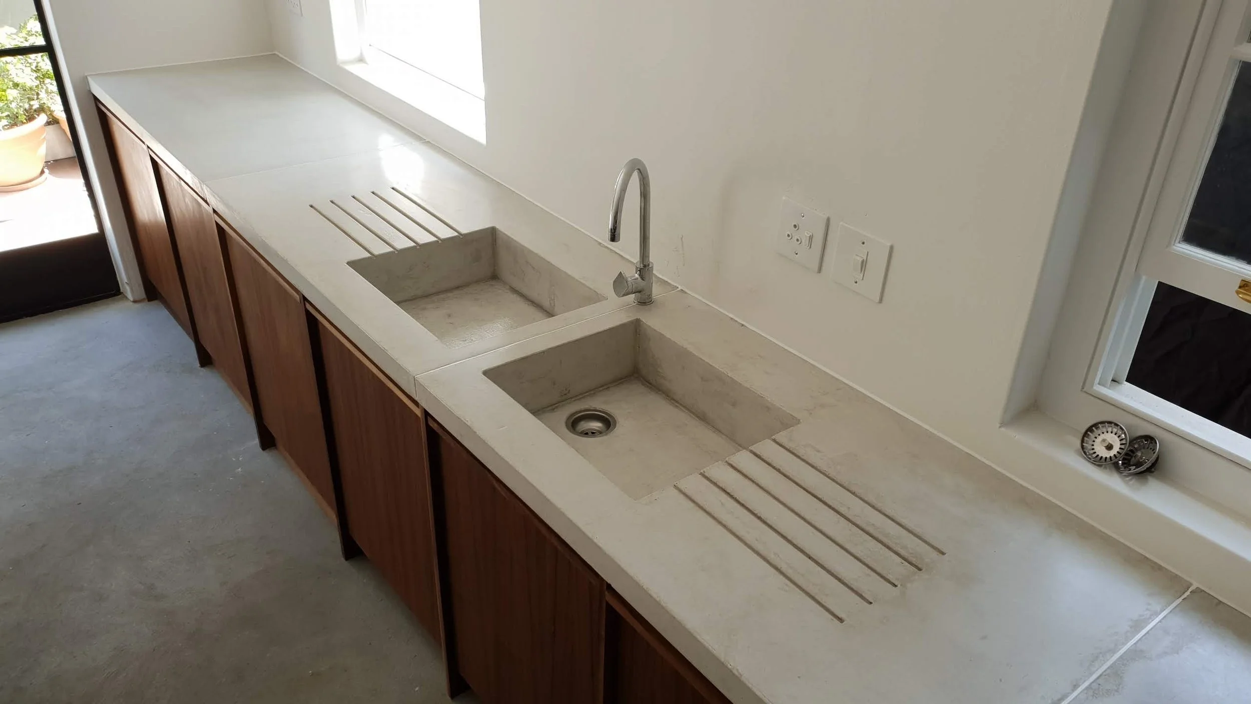 A kitchen countertop with dual sinks, one with a modern faucet, and drying racks on each side. There are electrical outlets and a window above the counter.
