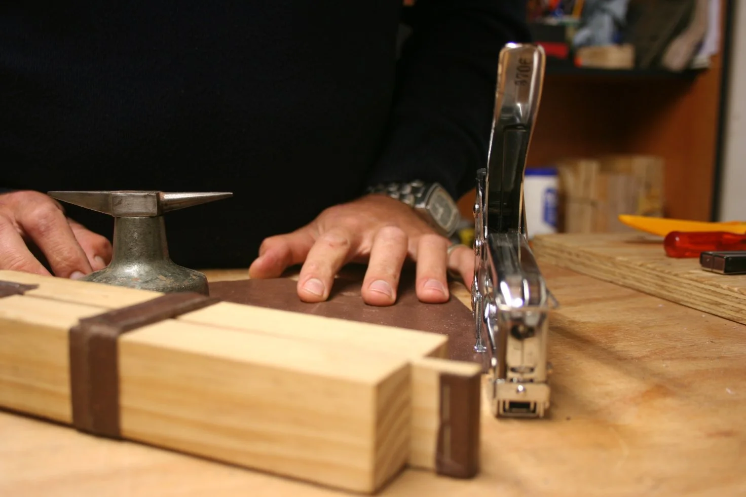 A person using a handheld electric planer on a wooden surface, with their other hand holding a metal clamp, in a woodworking workshop.