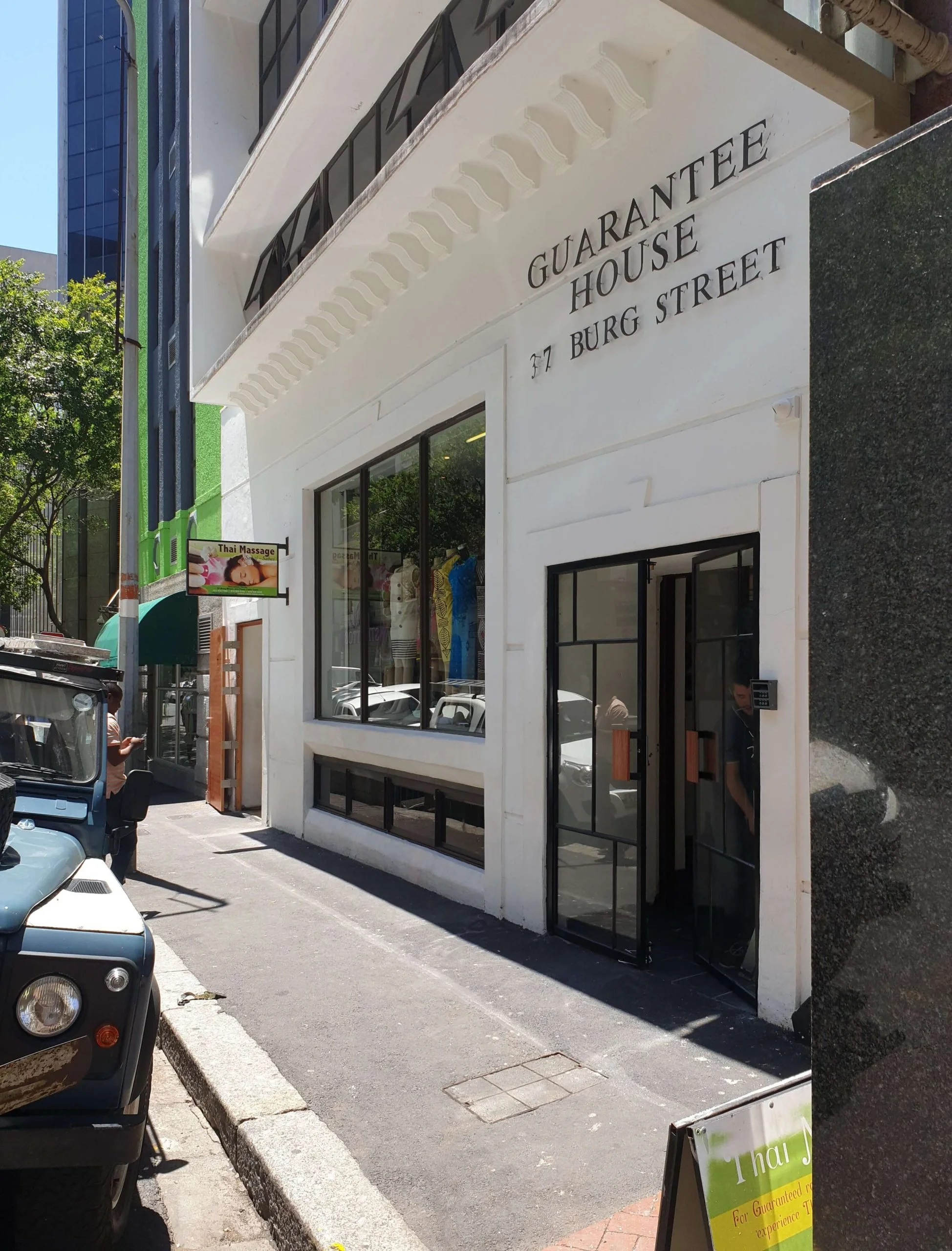 White building with sign reading "Guarantee House" and address "77 Burg Street" on a city sidewalk; large windows and a black door with a person partially visible inside.