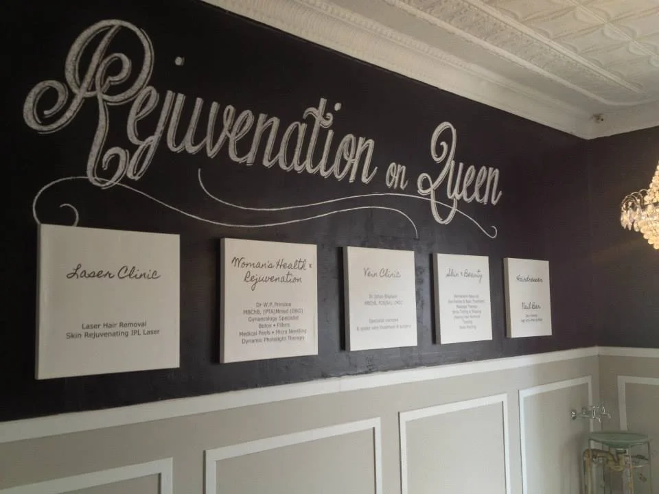 Interior wall with a gold chandelier, showcasing a blackboard-style sign that says "Rejuvenation on Queen" and five framed informational panels about beauty and skincare services.