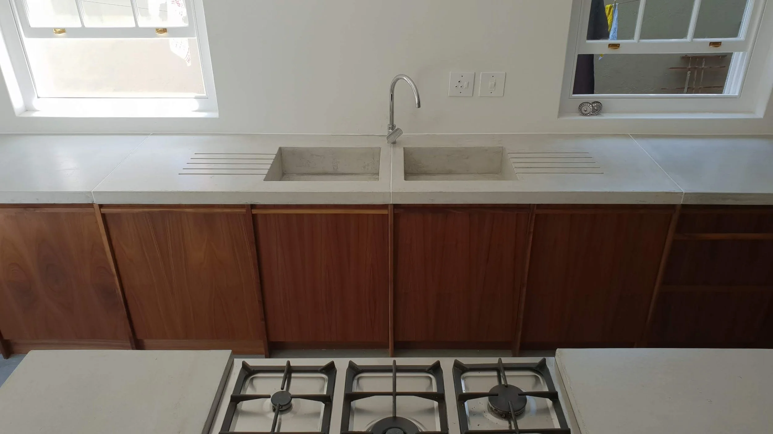 Kitchen sink with two basins and a faucet, countertop, wooden cabinetry, stove with three burners, two windows with sliding glass panes.