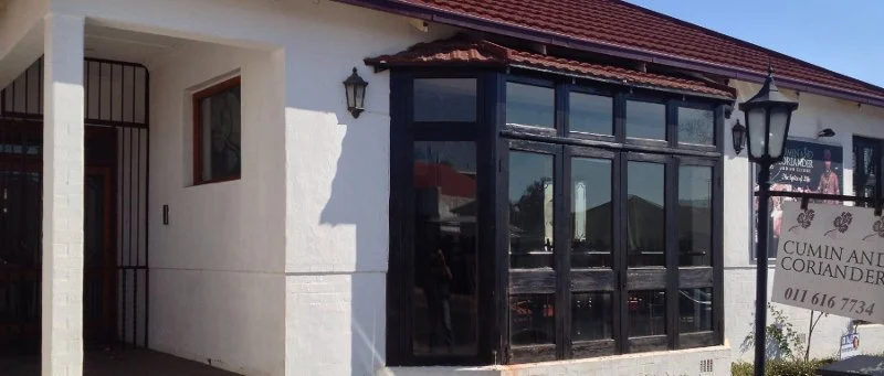 Exterior of a white building with a corner bay window, black trim, and lantern-style light fixtures, with a sign advertising cuisine, including cumin and coriander.