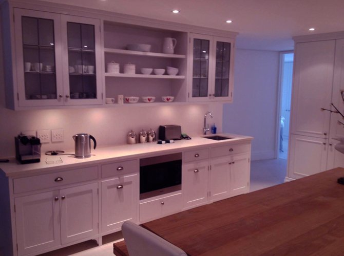 Modern kitchen with white cabinets, open shelves with bowls and dishes, and a wooden countertop table. Small appliances and utensils are on the counter. Walls have white paneling, and there are ceiling lights and a doorway leading outside.