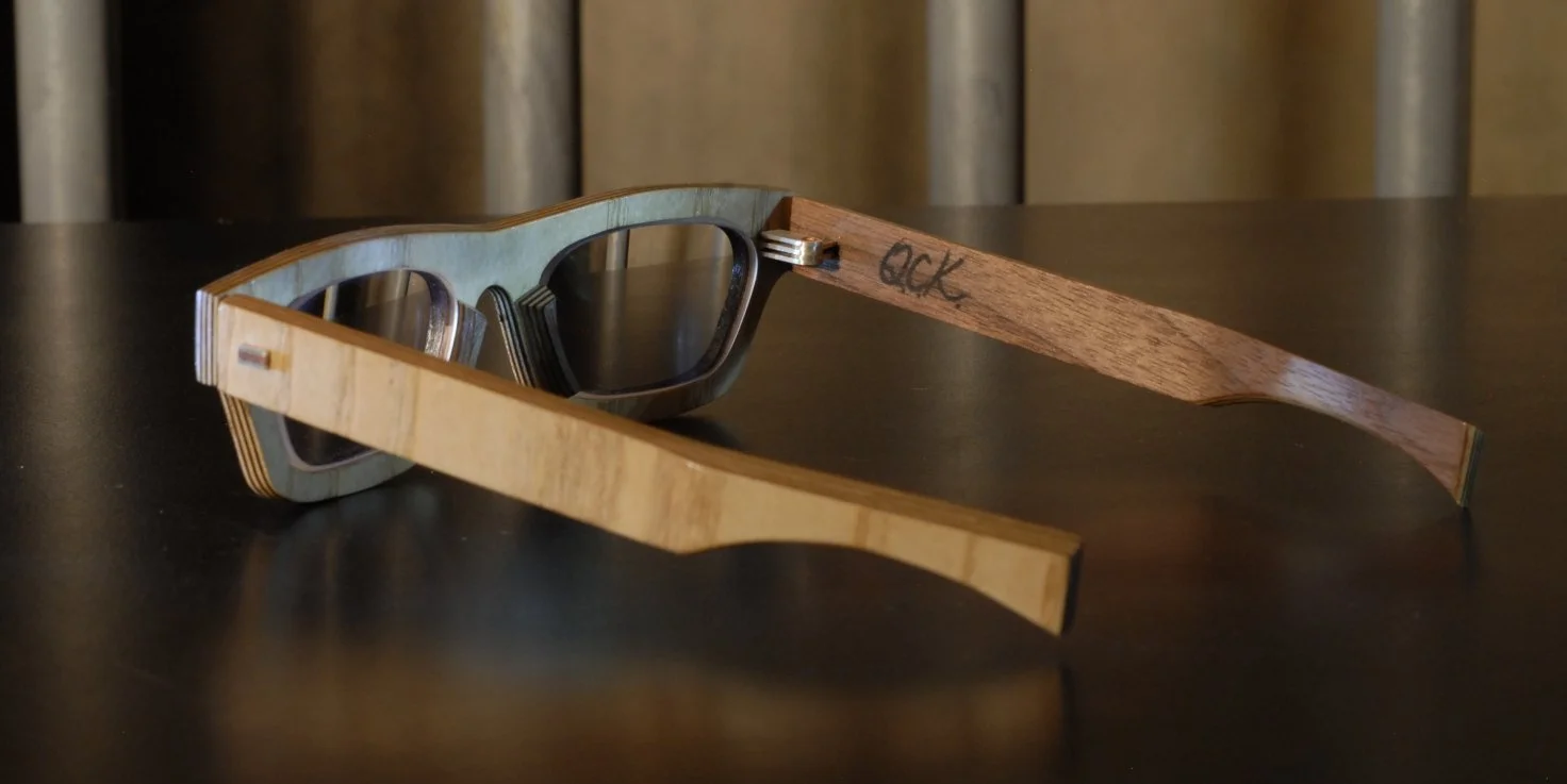 Pair of wooden sunglasses with dark lenses, one arm of the sunglasses has the word 'ock' written on it.